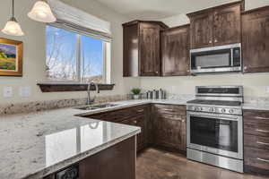 Kitchen featuring appliances with stainless steel finishes, light stone counters, pendant lighting, dark brown cabinetry, and dark wood finished floors