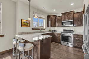 Kitchen with a peninsula, stainless steel appliances, a kitchen breakfast bar, decorative light fixtures, and light stone countertops