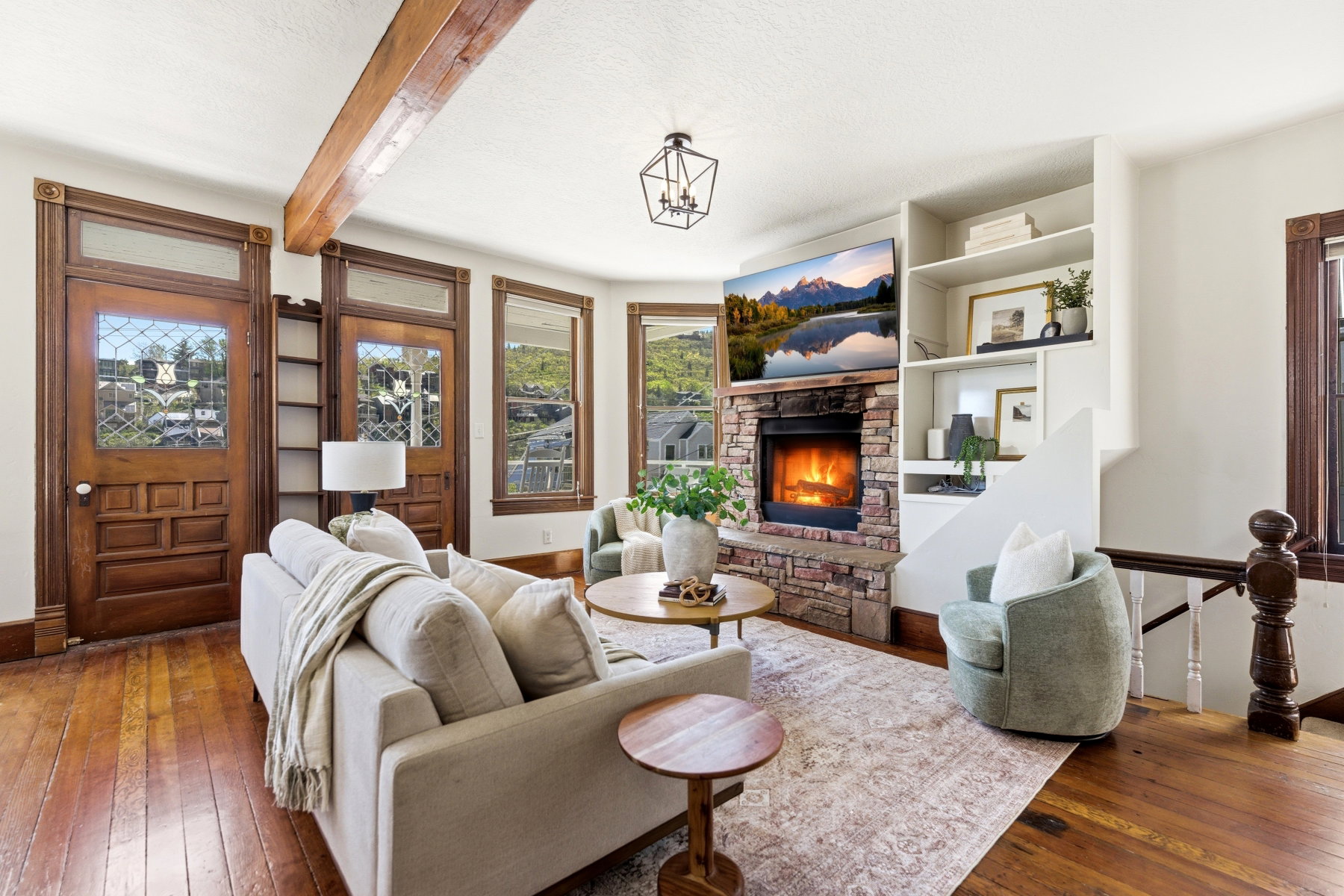 Living area featuring wood-finish flooring, a stone-clad fireplace with hearth, built-in shelving, a wood beam ceiling, and paneled wood doors with leaded glass inserts