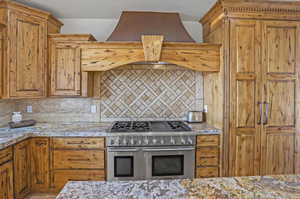 Kitchen with premium range hood, range with two ovens, tasteful backsplash, light stone countertops, and brown cabinetry
