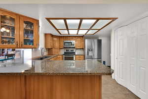 Kitchen with glass insert cabinets, stainless steel appliances, brown cabinetry, granite counters, and a peninsula