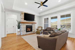 Living area with light wood-style flooring, a stone fireplace, a tray ceiling, a ceiling fan, and recessed lighting