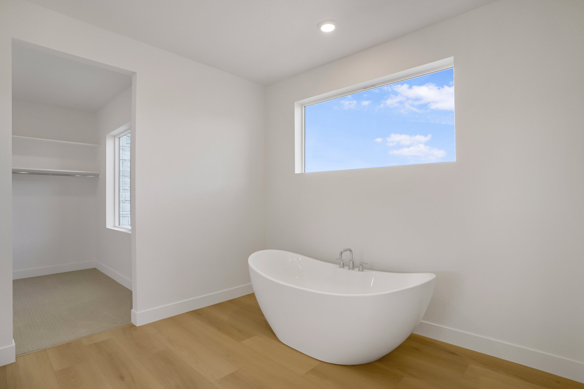 Freestanding soaking tub with polished chrome fixtures, horizontal window, wood-finish flooring, recessed lighting, and an adjacent walk-in closet with built-in shelving