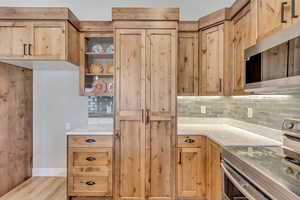 Kitchen featuring stainless steel appliances, glass insert cabinets, light stone counters, decorative backsplash, and light brown cabinets
