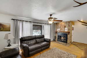 Living room featuring wood finished floors, a stone fireplace, a textured ceiling, and a ceiling fan