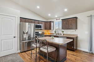 Kitchen featuring dark brown cabinetry, stainless steel appliances, a breakfast bar area, a kitchen island, and vaulted ceiling