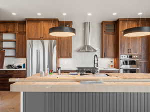 Kitchen featuring brown cabinets, stainless steel appliances, wall chimney exhaust hood, glass insert cabinets, and recessed lighting