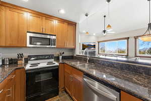 Kitchen featuring appliances with stainless steel finishes, vaulted ceiling, dark stone counters, brown cabinets, and pendant lighting