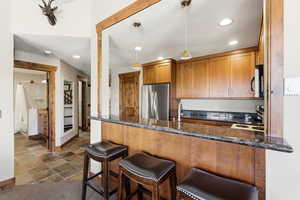 Kitchen with brown cabinetry, a peninsula, dark stone counters, appliances with stainless steel finishes, and recessed lighting