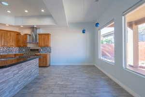 Kitchen featuring brown cabinetry, dark stone countertops, backsplash, decorative light fixtures, and wall chimney range hood