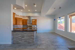 Kitchen featuring brown cabinets, pendant lighting, decorative backsplash, a peninsula, and wall chimney range hood