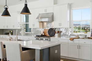Kitchen with white cabinetry, under cabinet range hood, decorative light fixtures, light stone counters, and a kitchen bar
