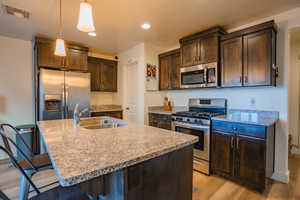 Kitchen featuring dark brown cabinetry, appliances with stainless steel finishes, light stone countertops, light wood-type flooring, and a center island with sink