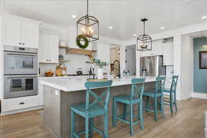 Kitchen featuring an island with sink, white cabinetry, appliances with stainless steel finishes, light wood-style flooring, and hanging light fixtures