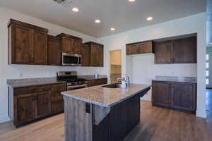 Kitchen featuring dark brown cabinets, stainless steel appliances, light stone counters, light wood-type flooring, and a kitchen island with sink