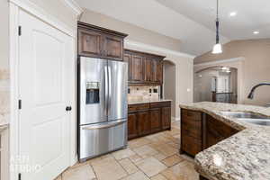Kitchen featuring arched walkways, dark brown cabinetry, stainless steel fridge, light stone countertops, and vaulted ceiling.