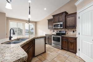 Kitchen with dark brown cabinets, appliances with stainless steel finishes, hanging light fixtures, lofted ceiling, and light granite countertops.