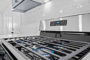 Kitchen view of stainless steel gas range, decorative backsplash, ventilation hood, and light stone countertops