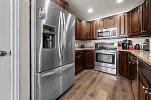 Kitchen featuring appliances with stainless steel finishes, dark brown cabinets, a textured ceiling, light stone countertops, and dark wood-style floors