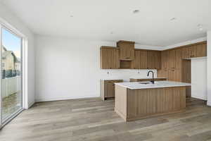 Kitchen with brown cabinetry, light wood finished floors, a kitchen island with sink, and light stone counters