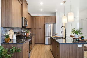 Kitchen featuring pendant lighting, stainless steel appliances, dark stone countertops, light wood-type flooring, and an island with sink
