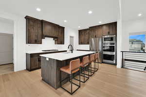 Kitchen featuring dark brown cabinets, a breakfast bar area, a kitchen island with sink, stainless steel appliances, and light wood finished floors