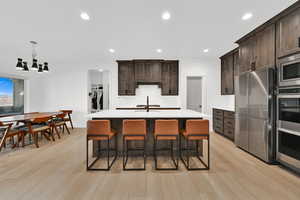Kitchen with dark brown cabinets, a breakfast bar area, appliances with stainless steel finishes, decorative light fixtures, and a center island with sink
