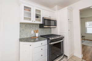 Kitchen featuring gas stove, glass insert cabinets, tasteful backsplash, dark stone counters, and white cabinetry