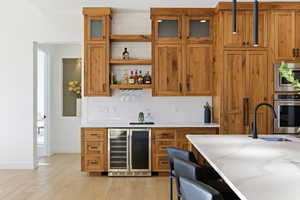 Indoor dry bar with brown cabinetry, wine cooler, backsplash, appliances with stainless steel finishes, and open shelves