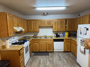 Kitchen featuring white appliances, light countertops, under cabinet range hood, brown cabinetry, and dark wood-type flooring