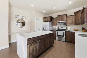 Kitchen featuring stainless steel appliances, light stone counters, dark brown cabinetry, a center island, and dark wood-style floors