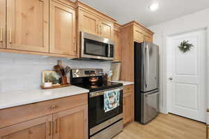 Kitchen featuring appliances with stainless steel finishes, light wood-style flooring, decorative backsplash, light brown cabinets, and light stone countertops