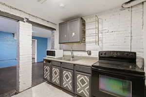 Kitchen with black electric range oven, brick wall, gray cabinetry, light countertops, and white microwave