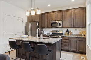 Kitchen featuring light stone countertops, dark brown cabinetry, stainless steel appliances, decorative light fixtures, and a kitchen island with sink