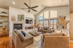 Living room featuring built in shelves, light wood-style floors, a glass covered fireplace, a ceiling fan, and high vaulted ceiling