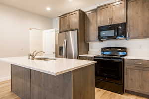 Kitchen with black appliances, an island with sink, light wood finished floors, recessed lighting, and light stone countertops