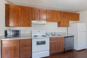 Kitchen with appliances with stainless steel finishes, brown cabinets, light countertops, dark wood-style floors, and under cabinet range hood