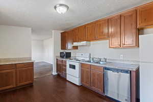 Kitchen with white appliances, a textured ceiling, light countertops, brown cabinets, and under cabinet range hood