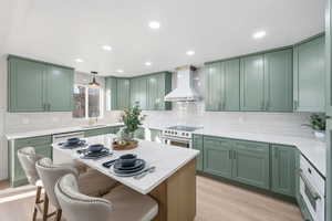 Kitchen featuring green cabinets, decorative light fixtures, wall chimney exhaust hood, a breakfast bar area, and light stone counters