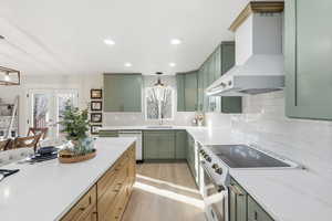 Kitchen featuring green cabinets, decorative light fixtures, wall chimney range hood, range with electric stovetop, and light stone counters