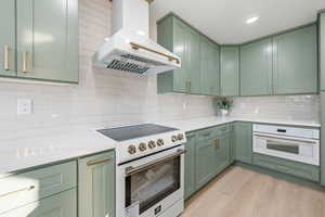 Kitchen with green cabinets, white appliances, wall chimney range hood, light stone countertops, and light wood-type flooring