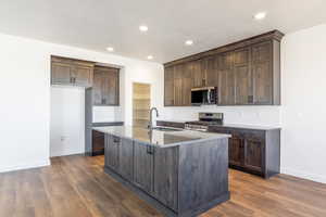 Kitchen featuring appliances with stainless steel finishes, dark brown cabinetry, a center island with sink, light stone counters, and recessed lighting