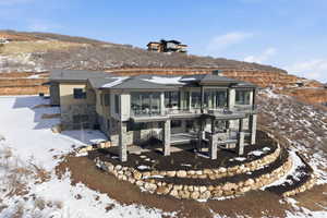 Snow covered back of property with a patio area, stone siding, a chimney, and a shingled roof