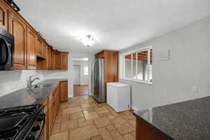 Kitchen featuring dark stone counters, black appliances, stone tile floors, wood finish cabinetry, and a textured ceiling