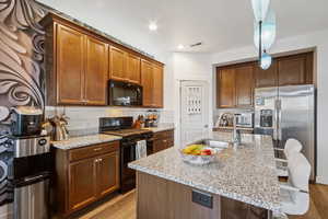 Kitchen with light stone countertops, black appliances, a breakfast bar, backsplash, and brown cabinets