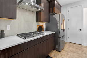 Kitchen featuring wall chimney exhaust hood, decorative backsplash, appliances with stainless steel finishes, light wood-style flooring, and dark brown cabinetry