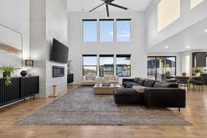 Living room with a tiled fireplace, ceiling fan, light wood finished floors, and a high ceiling