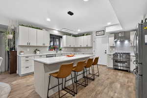 Kitchen featuring white cabinetry, appliances with stainless steel finishes, a kitchen breakfast bar, decorative light fixtures, and wall chimney range hood