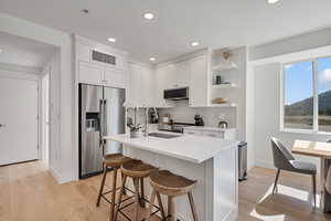 Kitchen with stainless steel appliances, white cabinetry, open shelves, an island with sink, and a breakfast bar