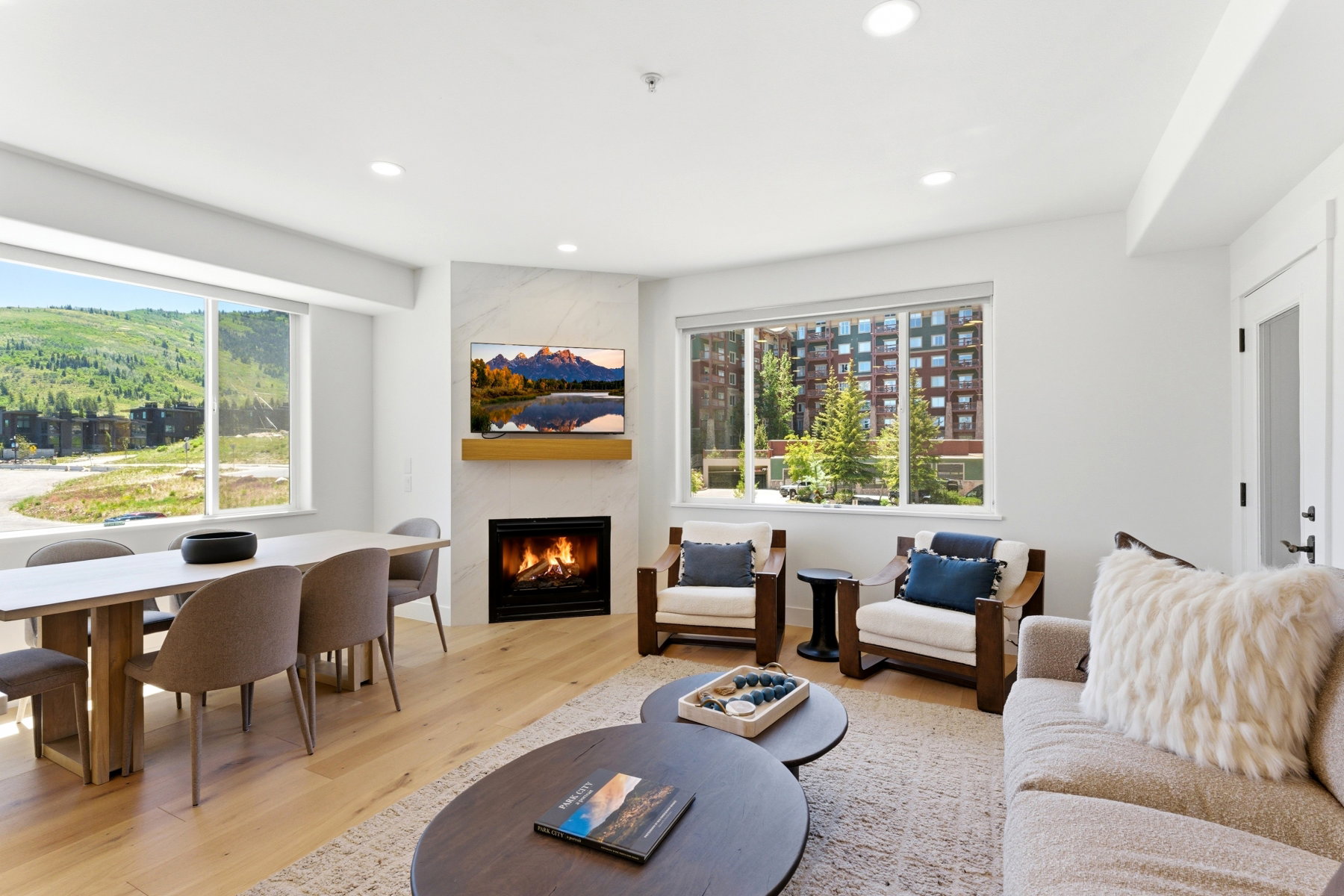 Open-concept living space with light wood-finish flooring, a contemporary fireplace with a stone surround and wood mantel, large windows, and recessed lighting
